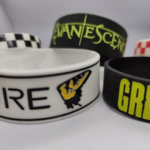 Könnte beinhalten: Eine Sammlung von Gummibändern in verschiedenen Designs. Ein Band ist weiß mit dem Wort "MORE" und einer Schmetterlingsgrafik. Andere Bänder sind schwarz mit neongrüner Schrift und Schachbrettmustern.