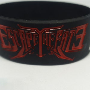 May include: Black rubber wristband featuring the red text "ESCAPE THE FATE". The text is in a stylized font and raised from the surface. The wristband is black with a glossy red finish to the text. A white accent is visible.