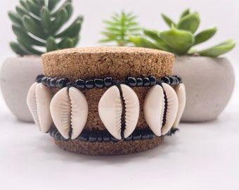 May include: A pair of bracelets featuring cream-colored cowrie shells and black beads. The bracelets are strung together with black cord. The bracelets are displayed on a cork surface, with green plants in the background.