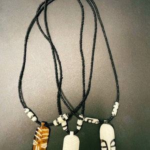 May include: Three necklaces with wooden and bone mask pendants. The necklaces are made of black beads and have gold accents. The masks are carved with intricate designs.