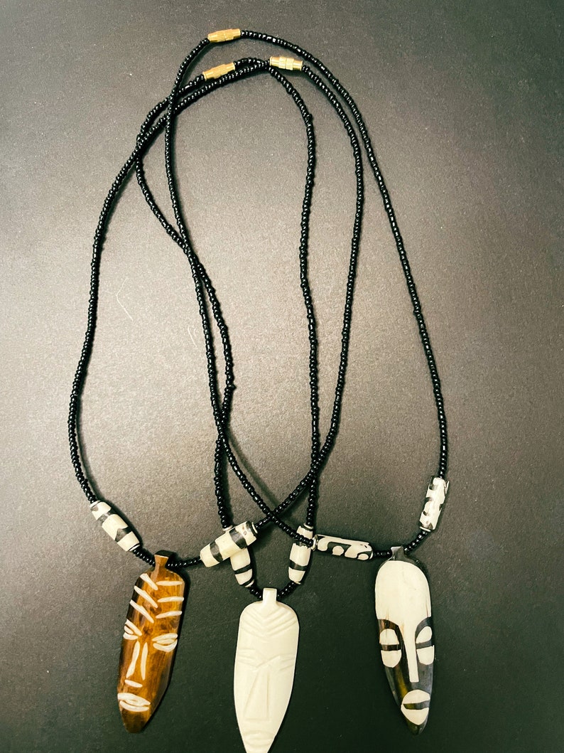 May include: Three necklaces with wooden and bone mask pendants. The necklaces are made of black beads and have gold accents. The masks are carved with intricate designs.