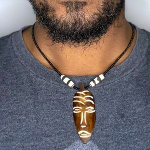 Può includere: Una collana di legno con un ciondolo a forma di maschera in legno intagliato. La maschera è marrone con dettagli bianchi e presenta un design tribale. La collana è composta da perline nere con accenti bianchi.