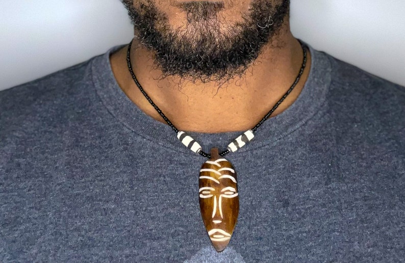 May include: A wooden necklace with a carved wooden mask pendant. The mask is brown with white details and has a tribal design. The necklace is made of black beads with white accents.