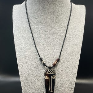 May include: A black beaded necklace with a wooden pendant featuring a carved tribal mask design. The mask is brown and black with white details.