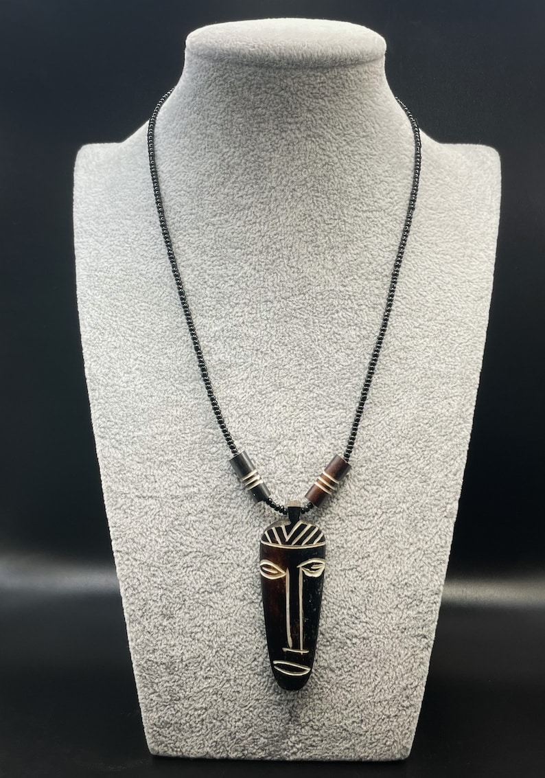 May include: A black beaded necklace with a wooden pendant featuring a carved tribal mask design. The mask is brown and black with white details.