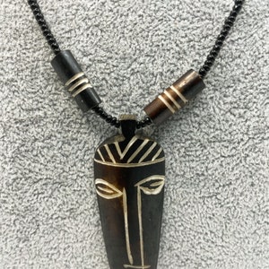 May include: A black beaded necklace with a wooden pendant featuring a carved tribal mask design. The mask is brown with white details and has a stylized face with eyes, nose, and mouth.