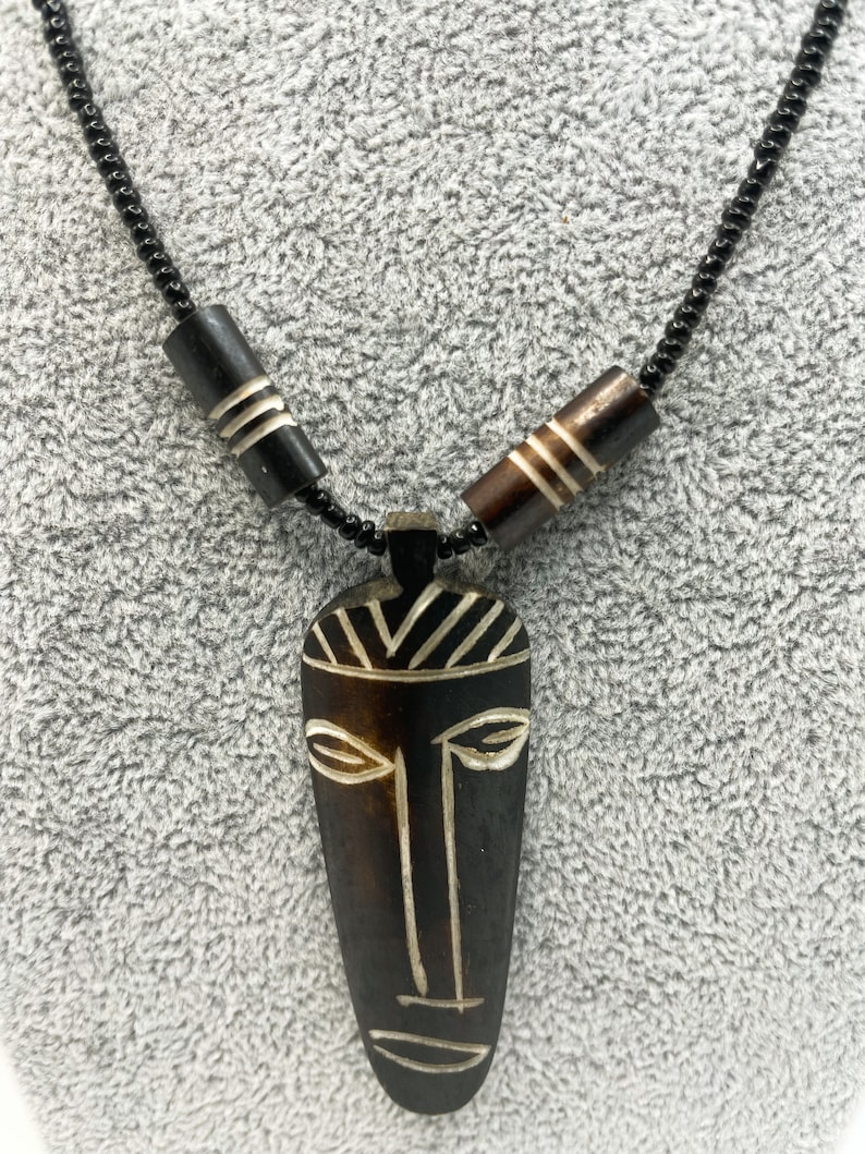 May include: A black beaded necklace with a wooden pendant featuring a carved tribal mask design. The mask is brown with white details and has a stylized face with eyes, nose, and mouth.