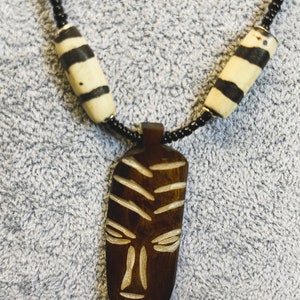 May include: A wooden necklace with a carved wooden mask pendant. The necklace is made of black beads and white and black striped beads.