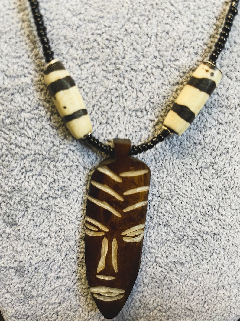 May include: A wooden necklace with a carved wooden mask pendant. The necklace is made of black beads and white and black striped beads.