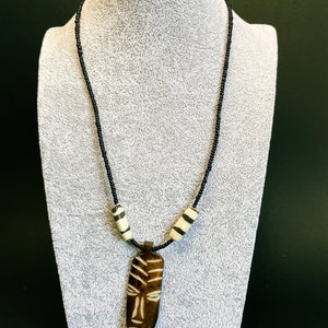 May include: A black beaded necklace with a wooden pendant carved with an African mask design. The pendant is brown with white details. The necklace also features two white and black striped beads.