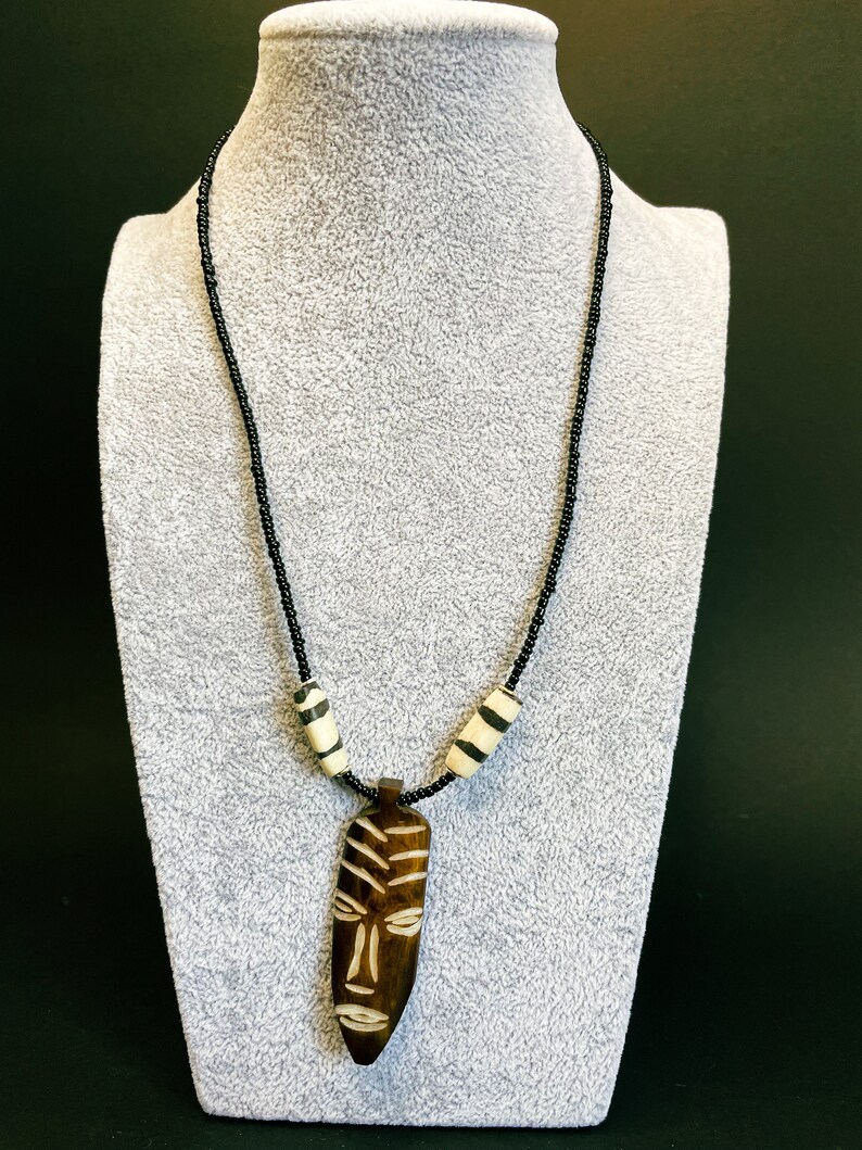 May include: A black beaded necklace with a wooden pendant carved with an African mask design. The pendant is brown with white details. The necklace also features two white and black striped beads.