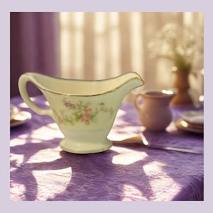 Puede incluir: Salsera de porcelana color crema con asa y pico, adornada con delicados motivos florales rosas y morados y ribete dorado. La salsera está sobre un mantel morado, con otros artículos de mesa al fondo.