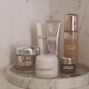 May include: A collection of La Mer skincare products on a white marble tray. The products include a face cream, eye cream, and a serum. The face cream jar is labelled "La Mer" and the eye cream jar is labelled "La Mer The Eye Concentrate". The serum bottle is labelled "La Mer". There is also a white jar labelled "La Peachy The Perfect Grinder".