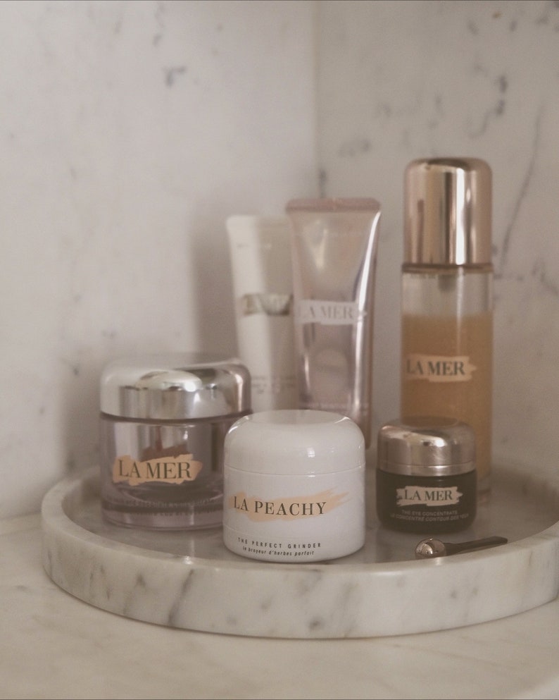 May include: A collection of La Mer skincare products on a white marble tray. The products include a face cream, eye cream, and a serum. The face cream jar is labelled "La Mer" and the eye cream jar is labelled "La Mer The Eye Concentrate". The serum bottle is labelled "La Mer". There is also a white jar labelled "La Peachy The Perfect Grinder".