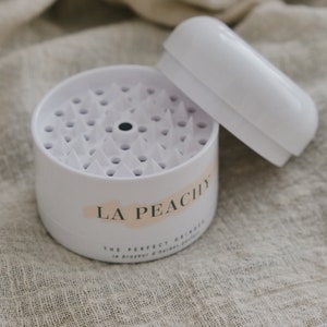 May include: White plastic container with a lid, labelled "LA PEACHY THE PERFECT GRINDER". The container has a circular, spiky surface inside.