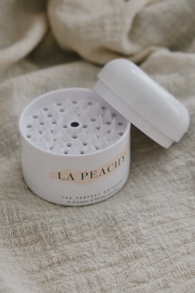May include: White plastic container with a lid, labelled "LA PEACHY THE PERFECT GRINDER". The container has a circular, spiky surface inside.