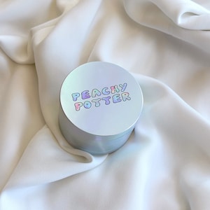 Peut inclure: Un récipient rond argenté avec une finition holographique et le texte "PEACHY POTTER" en lettres arc-en-ciel.