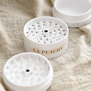 May include: White plastic grinder with a black centre and a label that reads "LA Peachy". The grinder has a textured surface with small, pointed teeth.