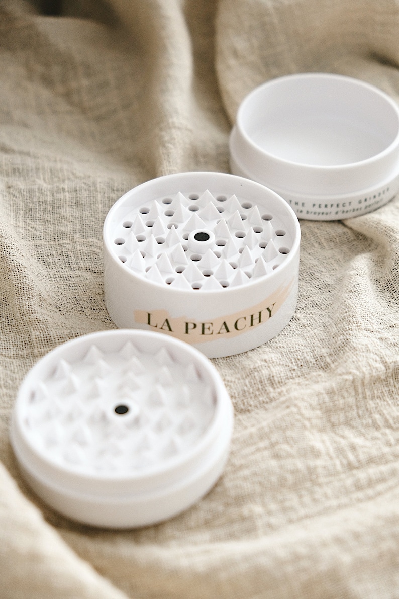 May include: White plastic grinder with a black centre and a label that reads "LA Peachy". The grinder has a textured surface with small, pointed teeth.