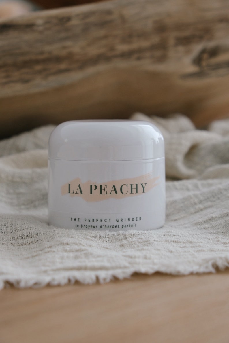 May include: White cylindrical grinder with a pink and white brushstroke design and the text "LA PEACHY THE PERFECT GRINDER le broyeur d'herbes parfait" in green.