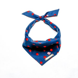 May include: A dark blue triangular bandana featuring a repeating pattern of small red apples. The fabric accessory includes a top tie closure, a single light pink apple-shaped button, and a rectangular gold-toned metal tag attached to the front.