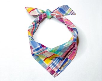 May include: A colorful plaid bandana with a gold metal tag. The bandana is tied in a knot at the top and has a blue, pink, yellow, and green plaid pattern.