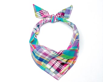 May include: A colorful plaid bandana with a pink, blue, green, and yellow pattern. The bandana is tied in a knot at the top and has a gold tag with the word "Bandana" on it.
