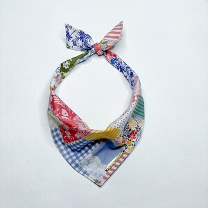 May include: A colorful patchwork bandana with a blue, red, green, yellow, and white floral pattern. The bandana is tied in a knot at the top and has a gold trim.