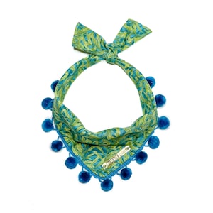 May include: A teal and green patterned dog bandana with a bow at the top and blue pom-poms along the bottom edge. The bandana has a tag with text on it.