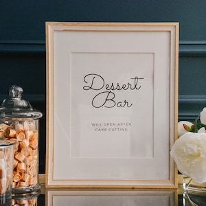 Pode incluir: Uma placa emoldurada com o texto "Dessert Bar" e as palavras "Will open after cake cutting" em letras pretas sobre fundo branco. Dois frascos de vidro cheios de caramelos dourados estão em primeiro plano.