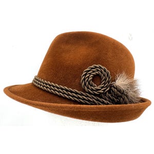 Puede incluir: Un sombrero de fieltro marrón con una banda decorativa de cuerda trenzada que rodea la copa. La banda presenta un detalle de nudo de cuerda circular en el lateral, acentuado con un pequeño mechón de pelo marrón claro detrás del adorno.