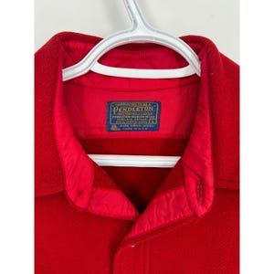 May include: Close-up of a red wool jacket with a quilted lining and a button closure. The jacket features a label with the brand name "Pendleton" and details about the wool composition. The jacket is hanging on a white hanger.