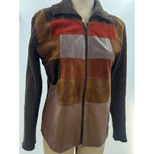 May include: A zip-up jacket featuring a patchwork design of suede and leather panels in shades of brown, rust, and tan. The garment includes long, dark brown ribbed knit sleeves and a matching ribbed knit collar, with a central vertical zipper closure.