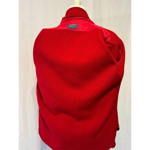 May include: A bright red fleece jacket with a high collar and a small blue and white label on the back. The jacket is displayed on a mannequin, showcasing its full shape and design. The jacket's material appears soft and warm.