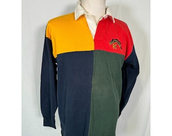 May include: A long-sleeved rugby shirt with a multi-coloured design. The shirt is yellow, red, dark blue, and green. The shirt has a white collar and a button placket. The shirt has the text "Whistler BC" embroidered on the front.
