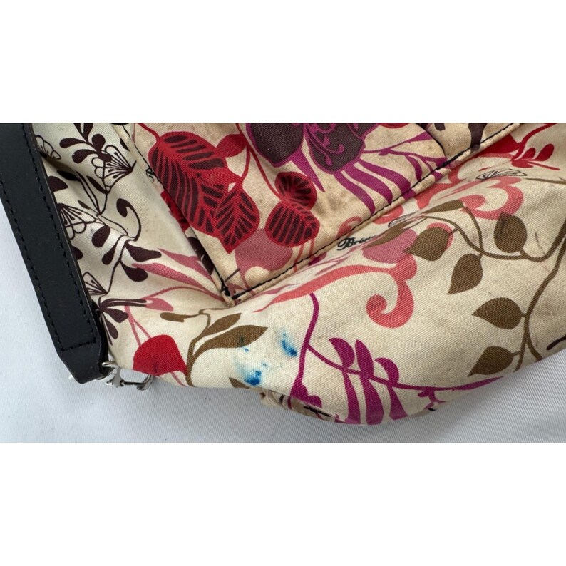 May include: A section of a floral patterned handbag interior. The fabric features a cream background with burgundy, pink, and purple floral designs. A black leather strap is visible on the left side. The bag appears to be used.