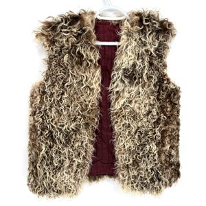 Puede incluir: Un chaleco mullido y abierto con un exterior rizado y texturizado en tonos marrones y crema. El forro interior es de color burdeos intenso. El chaleco está colgado de una percha blanca sobre un fondo blanco.