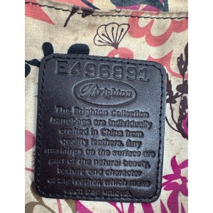 May include: Close-up of a dark brown leather tag with the Brighton logo and the number E496891. The tag is set against a floral patterned fabric with pink and cream tones. The text on the tag reads "The Brighton Collection handbags are individually crafted in China from quality leathers."
