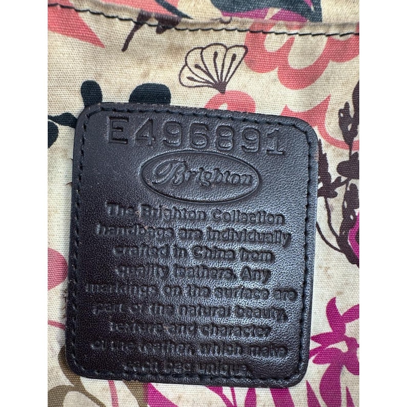 May include: Close-up of a dark brown leather tag with the Brighton logo and the number E496891. The tag is set against a floral patterned fabric with pink and cream tones. The text on the tag reads "The Brighton Collection handbags are individually crafted in China from quality leathers."