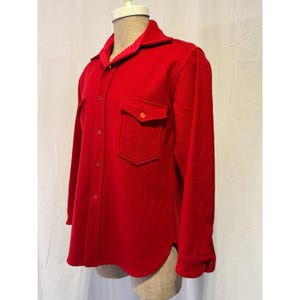 May include: A bright red wool shirt jacket with two front pockets and a button-down closure. The jacket has a collar and long sleeves. The interior collar lining is a contrasting red fabric. The jacket is displayed on a mannequin.