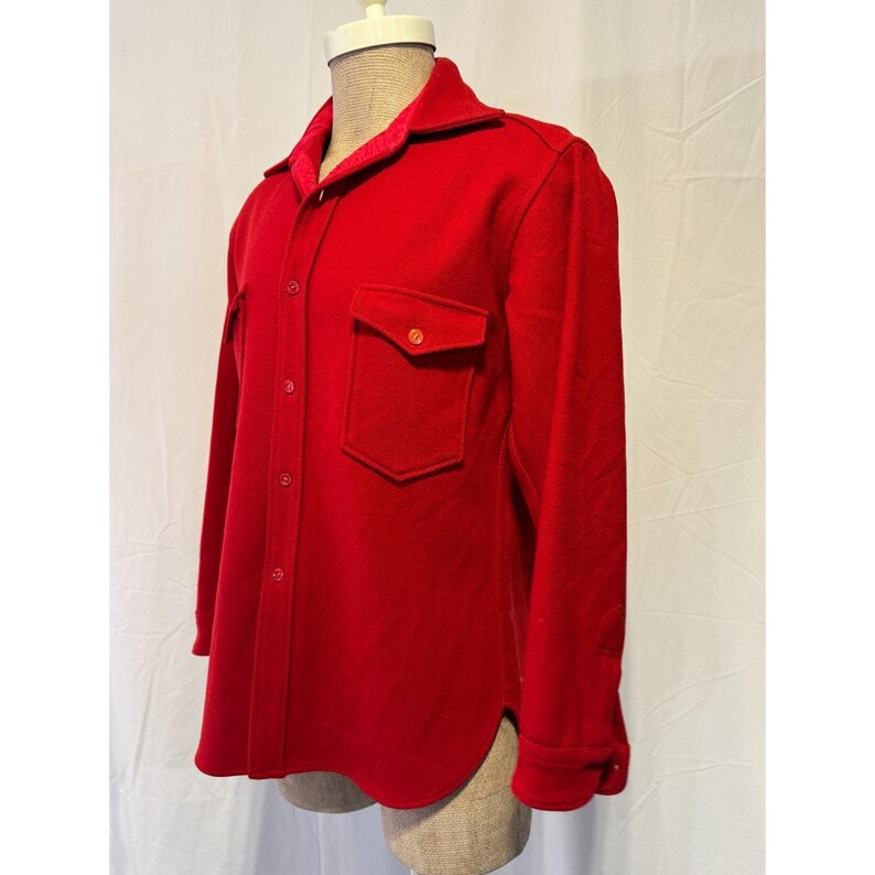 May include: A bright red wool shirt jacket with two front pockets and a button-down closure. The jacket has a collar and long sleeves. The interior collar lining is a contrasting red fabric. The jacket is displayed on a mannequin.