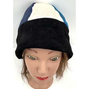 May include: A multi-colored beanie featuring a wide black band at the base, with vertical panels of navy blue, cream, and teal extending from the top. The hat is constructed from a soft, suede-like material with a rounded crown and a fitted shape.