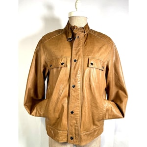 May include: A tan leather jacket with a button-up front, two chest pockets, and a collared neckline. The jacket has a classic design with a buckle detail at the collar and is displayed on a mannequin.