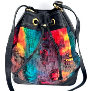May include: A black leather bucket bag with a colorful, textured fabric body. The fabric features a vibrant rainbow of colors, including red, orange, yellow, green, and blue. The bag has a drawstring closure and gold-tone hardware.