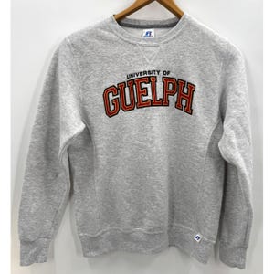 May include: A light grey crewneck sweatshirt featuring the text UNIVERSITY OF GUELPH in red and black block lettering across the chest. The garment has long sleeves, a ribbed collar with a V-stitch detail, and a small Russell brand logo tag.