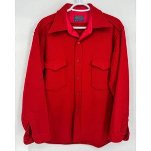 May include: A vibrant red wool shirt featuring two chest pockets and a button-down design. The shirt has a broad collar and long sleeves with buttoned cuffs. A small label is visible at the collar. The shirt is suspended on a white hanger.