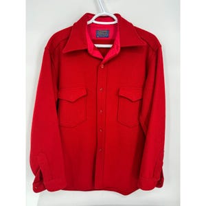 May include: A bright red wool shirt jacket with a collared neckline, two chest pockets, and button closures. The jacket is hanging on a white hanger. A small label is visible on the inside collar.