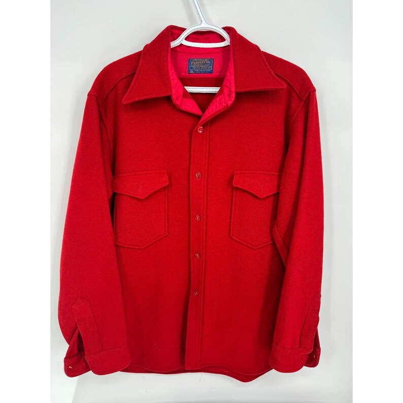 May include: A bright red wool shirt jacket with a collared neckline, two chest pockets, and button closures. The jacket is hanging on a white hanger. A small label is visible on the inside collar.