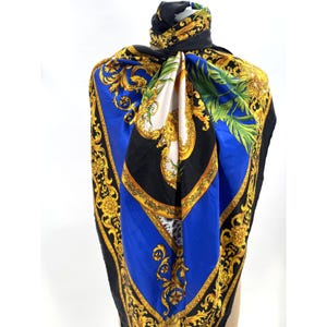May include: A square scarf featuring a vibrant blue, black and white colour palette with intricate gold filigree, scrollwork and floral patterns. The design includes green palm leaf motifs and a central white panel, all finished with a black border.