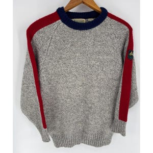 May include: A gray knit sweater with red stripes on the shoulders and sleeves. The sweater has a navy blue collar and a small emblem on the left sleeve. The sweater is displayed on a wooden hanger.